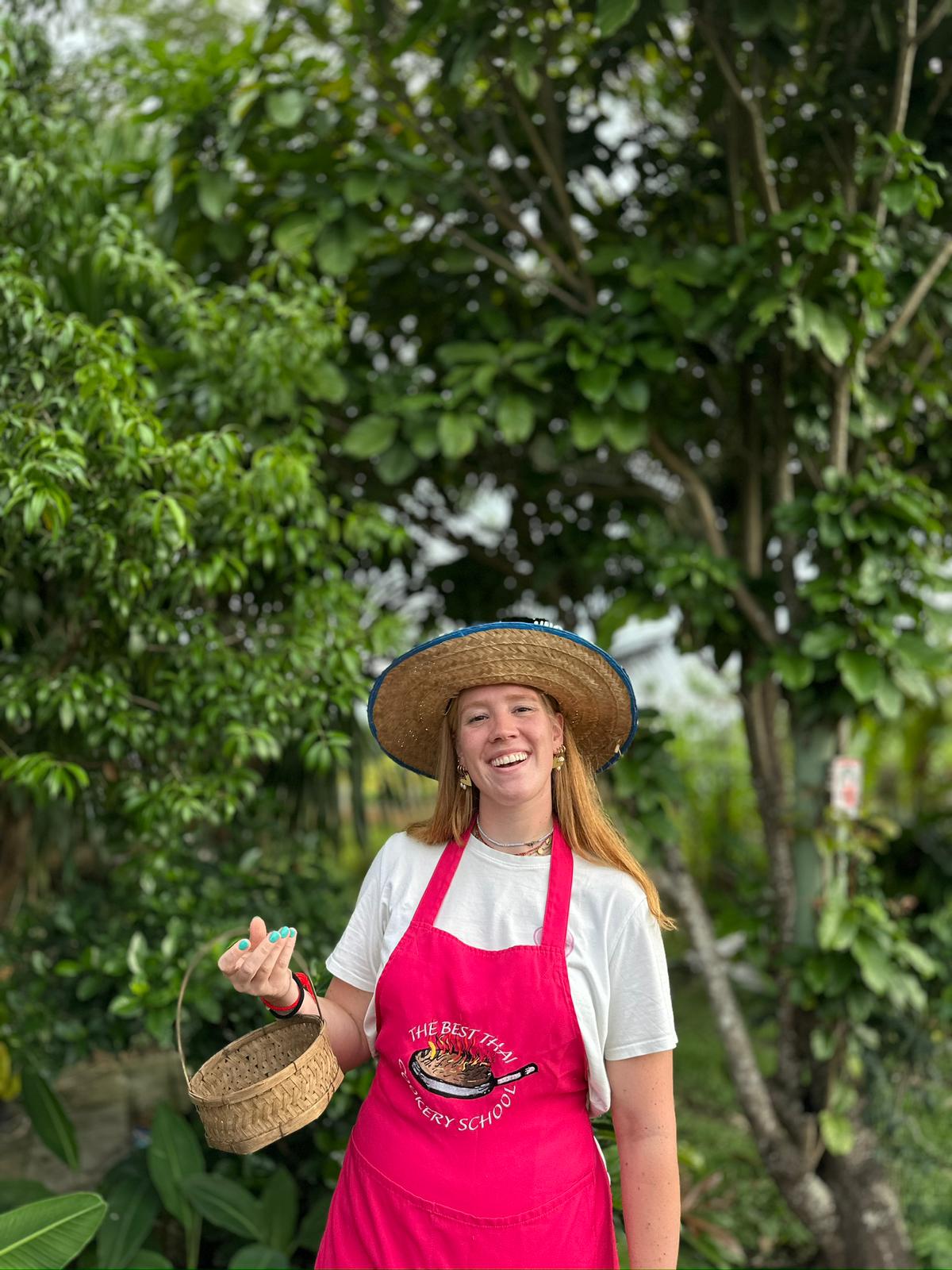 Student in the organic farm garden wearing a The Best Thai Cookery School apron and straw hat, holding a wicker basket